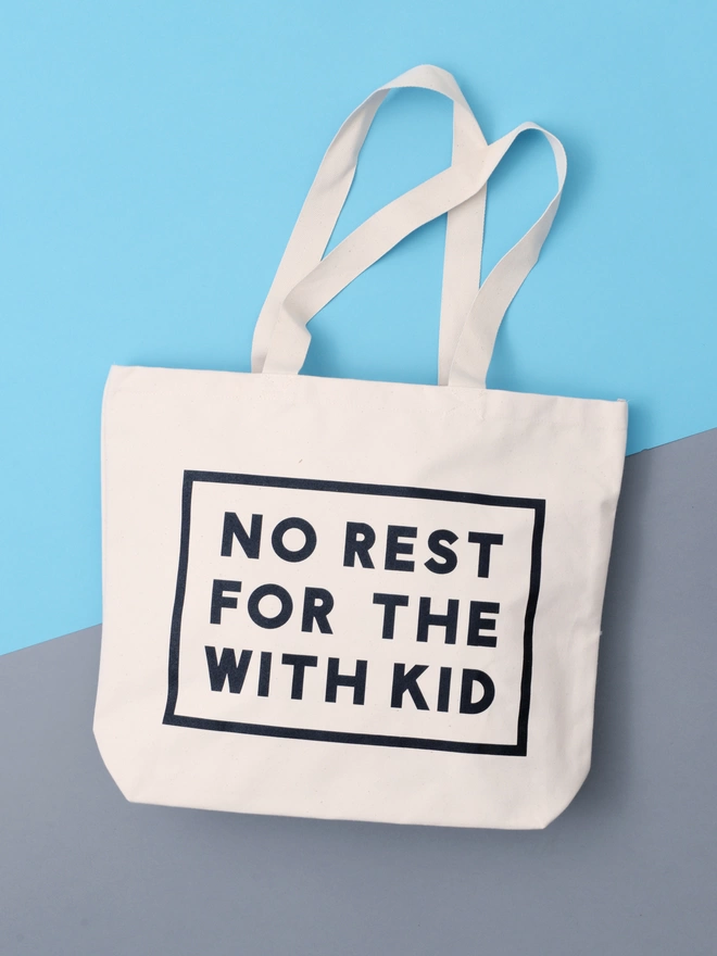 a large natural canvas tote bag bearing the words no rest for the with kid laying on a blue and grey backdrop