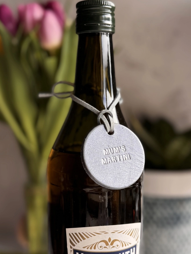 A dark glass bottle with a pewter leather tag hanging around the neck by a leather cord. The tag is hand-stamped with 'MUM'S MARTINI' in gold lettering, with pink tulips and a rustic background. A perfect personalised Mother’s Day gift or home bar accessory