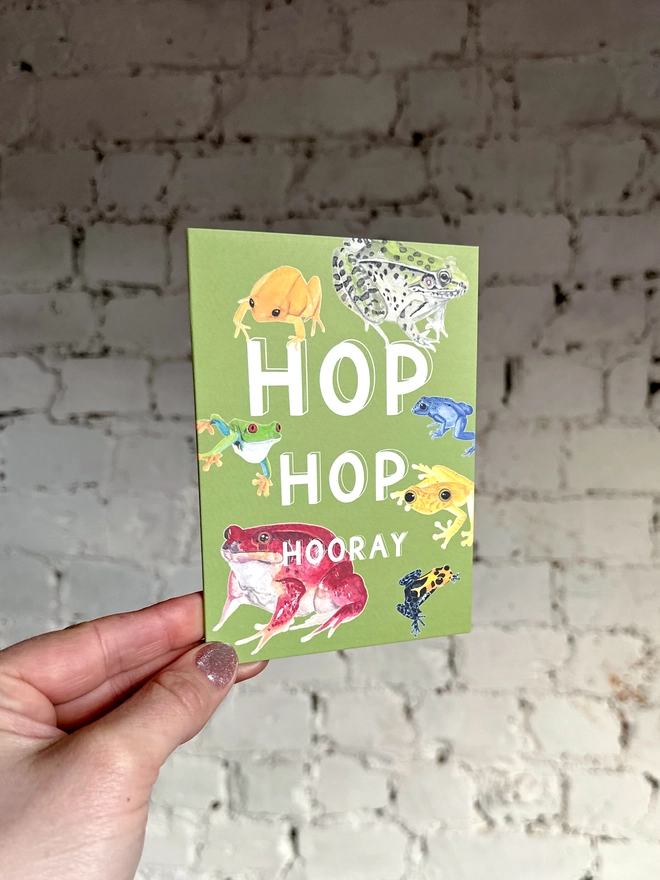 a green greetings card featuring a selection of frog illustrations with the text “hop hop hooray”