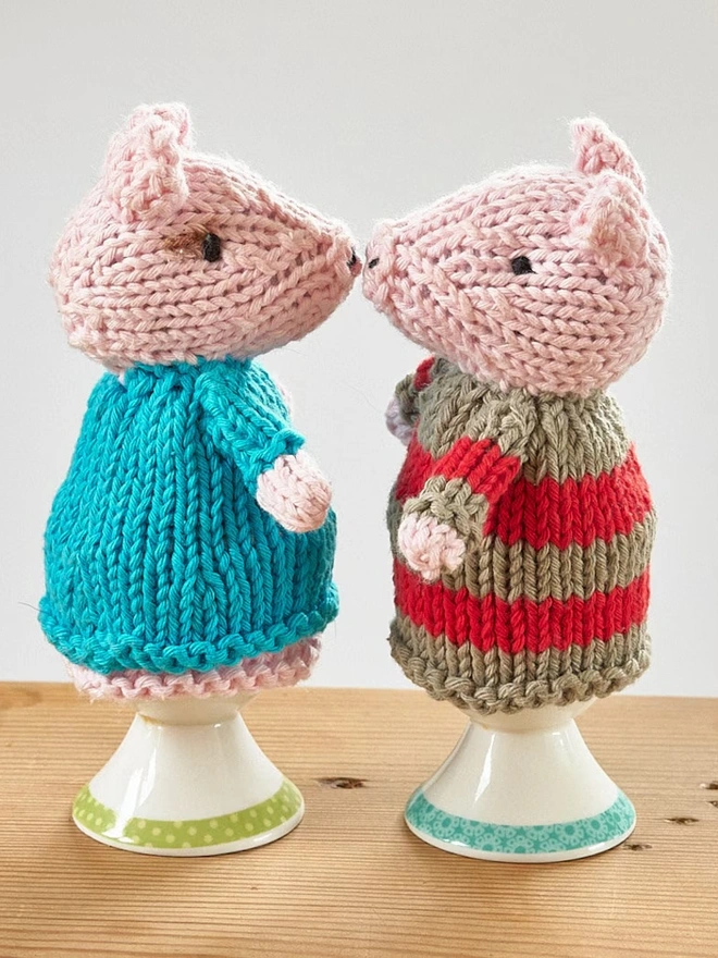 Egg Cosy Pigs Table Decoration (Set Of 2), a pair of colourful pig cosies sitting on a wooden surface. 