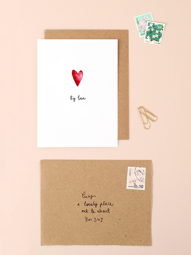 An image of a mini, rectangular white greetings card with an illustration of a red heart with a hand written 'Big love' message in black ink seen under the central illustration. The card is seen on top of a brown rectangular kraft envelope.