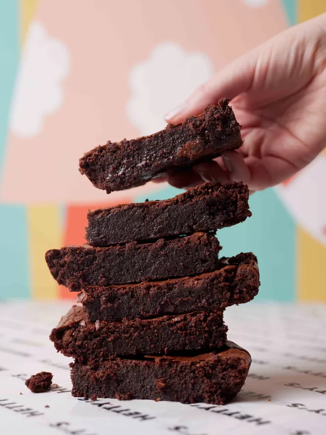 Gluten free classic fudge brownies in a stack with a hand adding another brownie to the top of the stack against a colourful background
