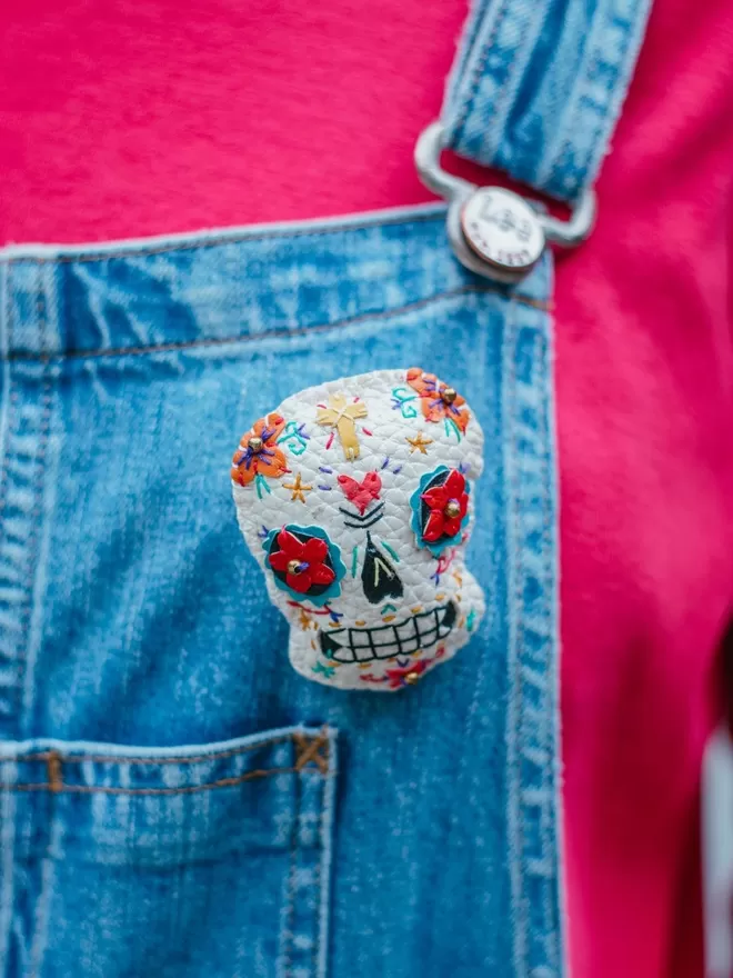 A hand stitched faux leather white Mexican Day of the Dead inspired sugar skull brooch with  colourful stitching and gold bead details, on a wooden background with red roses