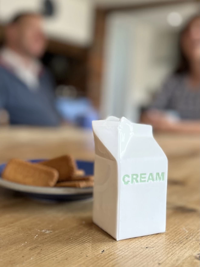 A handmade ceramic jug that emulates a paper carton, has ‘cream’ recessed in green on its side.