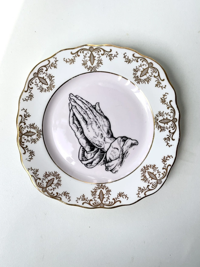 a vintage ornate plate that has gold and white patterned border and a pink centre, and in the centre i have print a black and white illustration of praying hands in a vintage style