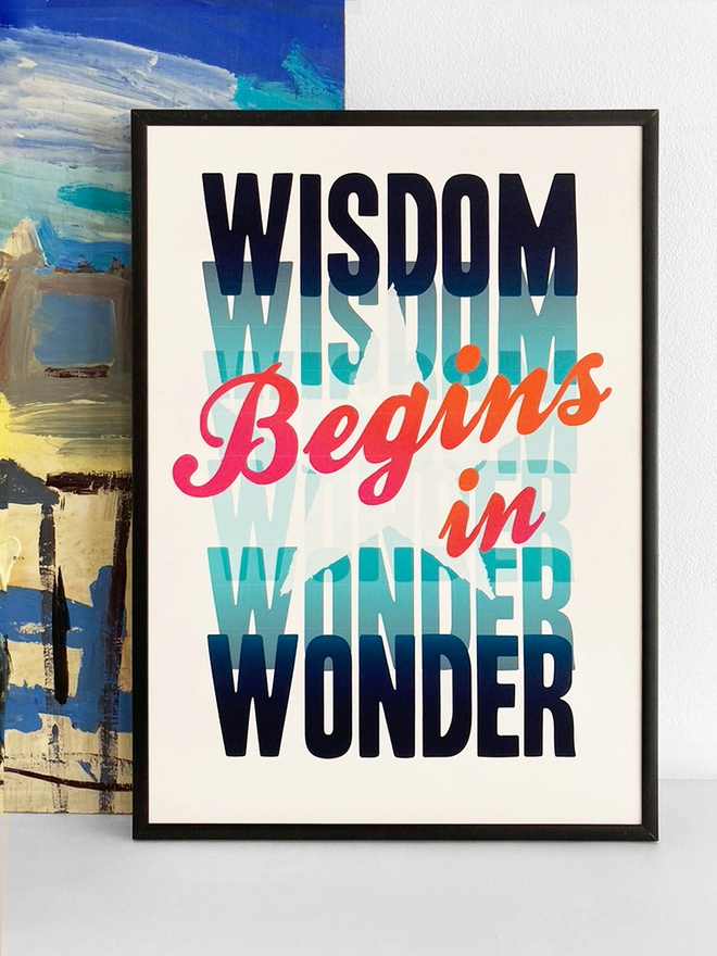 Framed multicoloured typographic print of Socrate’s famous quote - “Wisdom Begins In Wonder”  The print rests against a blue and yellow abstract painting.