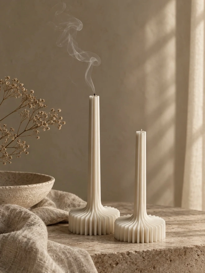 Pair Of White Flair Candles (Set Of 2). Two cream sculptural candles on a travertine surface, one just extinguished with a curl of smoke, beside a stone bowl, linen and dried gypsophila. The calm, minimalist design adds a serene, spa-like feel to any room.