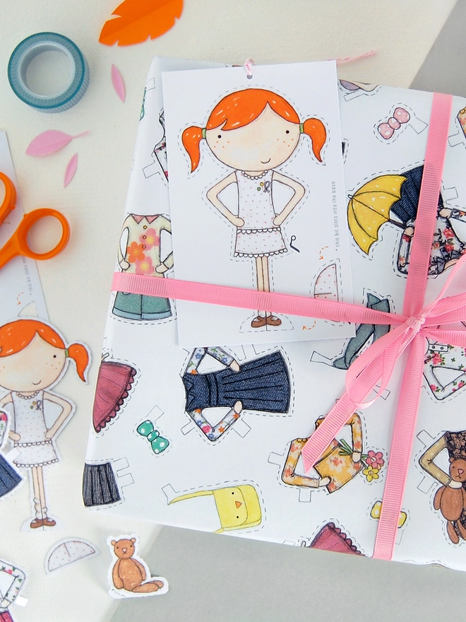 A gift wrapped in wrapping paper with illustrated outfits for the included paper doll tag.