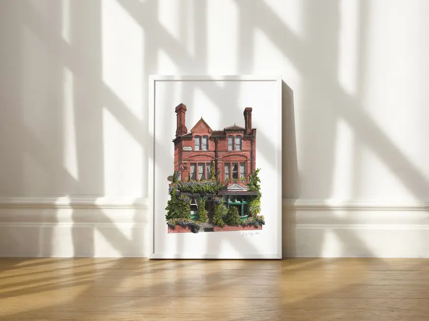 Nunhead Skehans Giclée Print (Unframed), a print of a brick pub in a white frame against a plain backdrop on a wooden surface. 