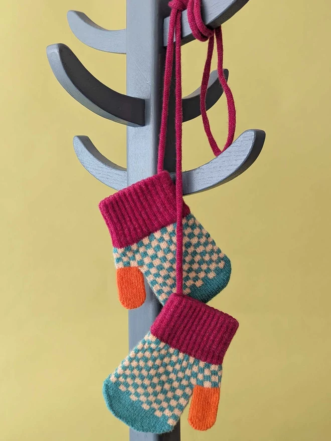 Kids Green And Peach Check Mittens On A String Hanging On A Coat Stand In Front Of  A Green Background