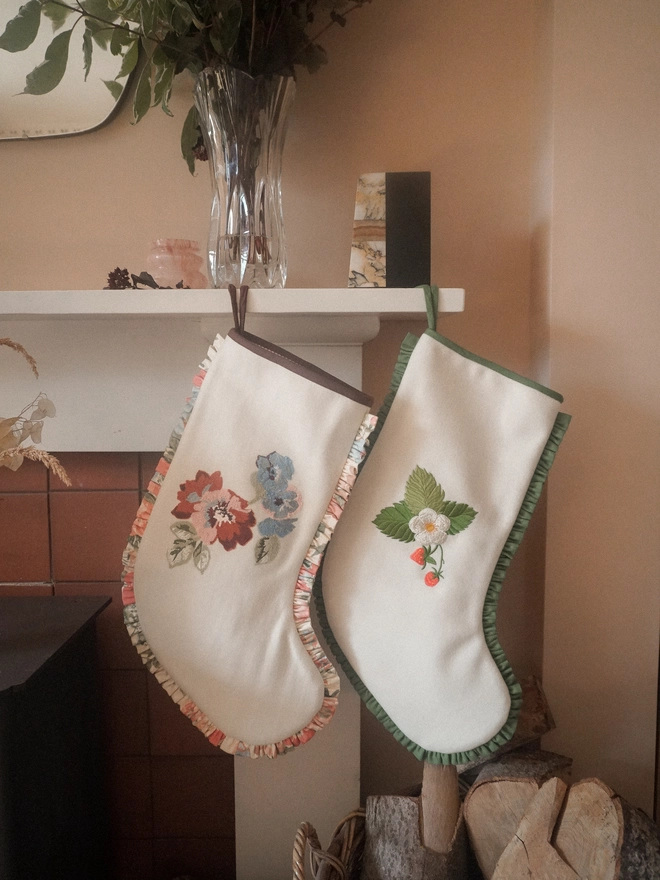 Two handmade ivory Christmas stockings hanging from a fireplace, embroidered with colourful floral and strawberry designs and finished with gathered fabric trims.