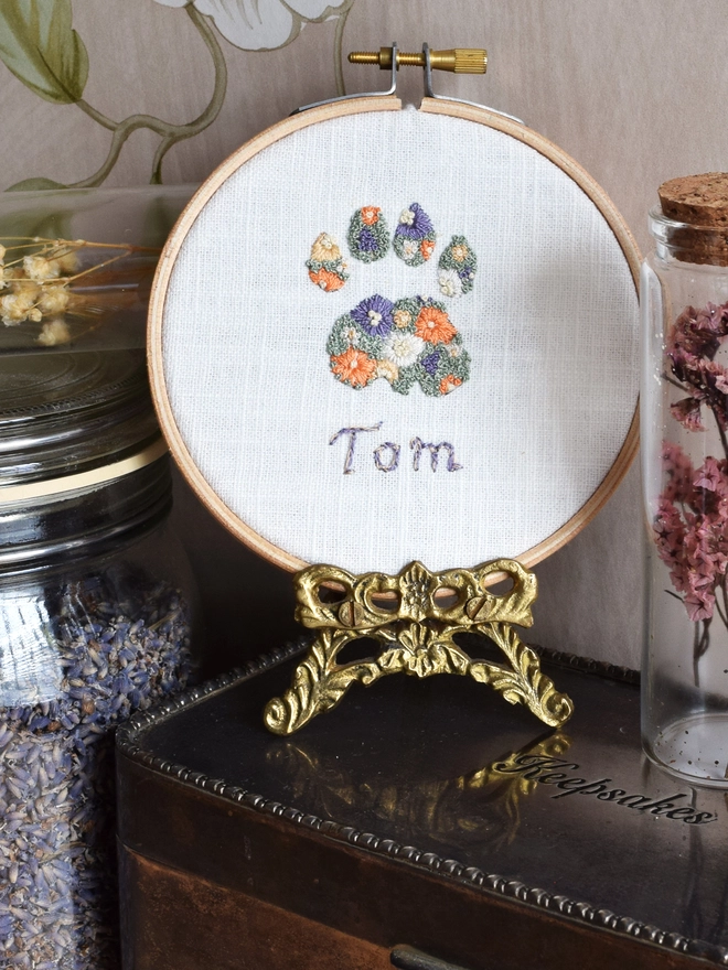 An embroidered Sunshine Garden Cats Paw, of Golden Yellows and Bright Orange Blossoms with Green French Knot grass background.  Displayed in a hoop frame on a gold metal stand on top of a silver keepsake box.