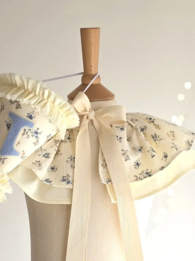 Vintage-inspired floral birthday cape and party hat set in soft vanilla cotton with delicate blue ditsy flowers.