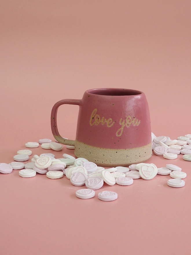 a bright pink handmade stoneware mug, with the saying love you on the front, sits on a pink background surrounded by love heart sweets