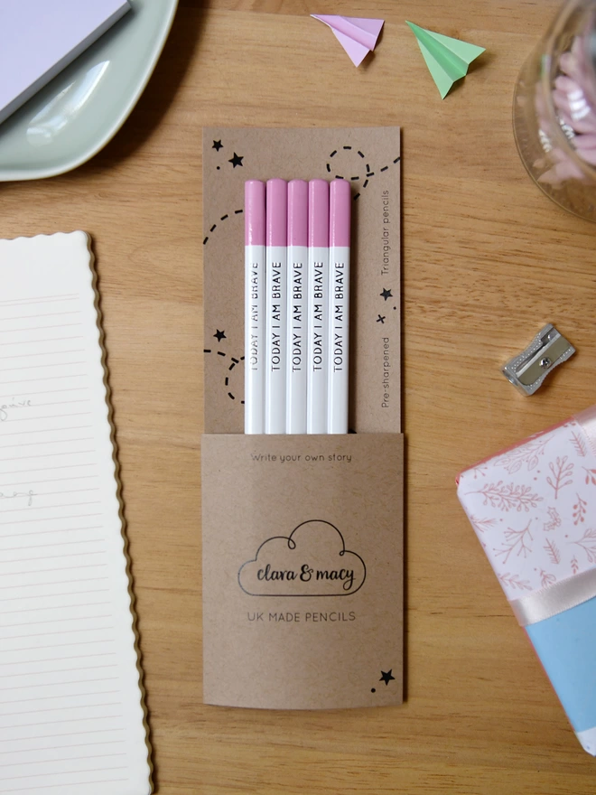 Five white pencils with pink tips and the words Today I Am Brave written along the side, sit within cardboard packaging on a wooden desk with stationery items.