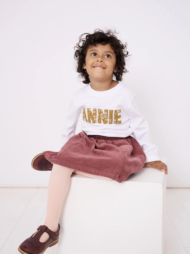 A white long sleeve cotton t-shirt appliquéd with the name Annie in a floral Liberty print, worn by a 3 year old girl
