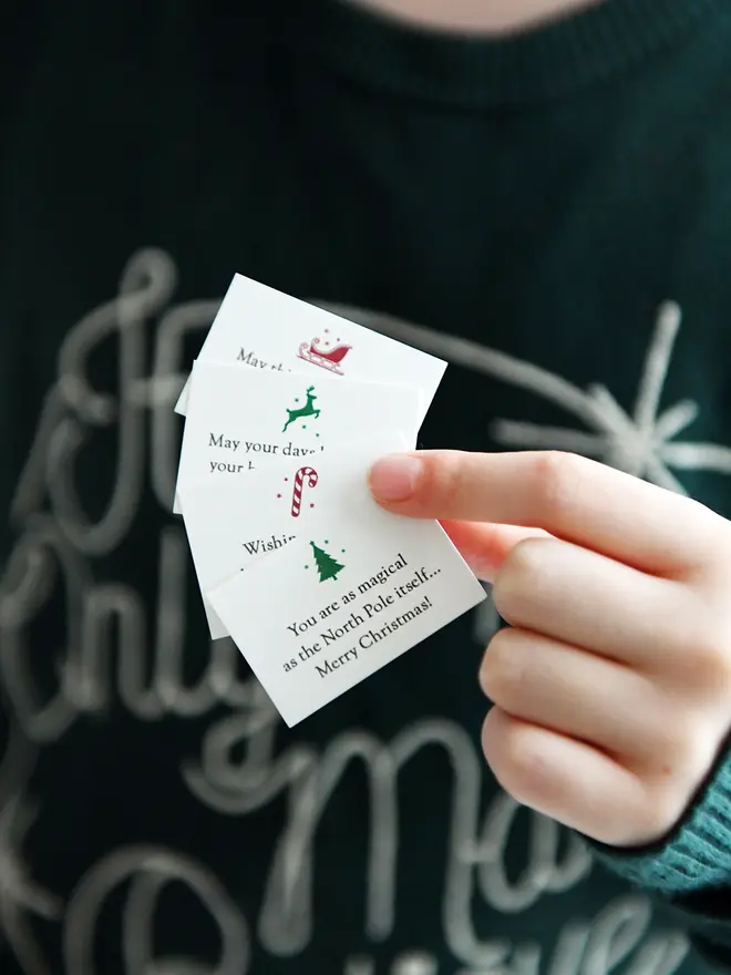 A child wearing a deep green jumper is holding tiny Christmas wish cards with festive icons, perfect as DIY Christmas cracker fillers or stocking stuffers.