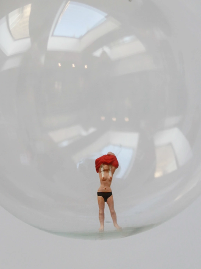 Miniature figure of woman playfully pulling off her top in a glass Christmas bauble