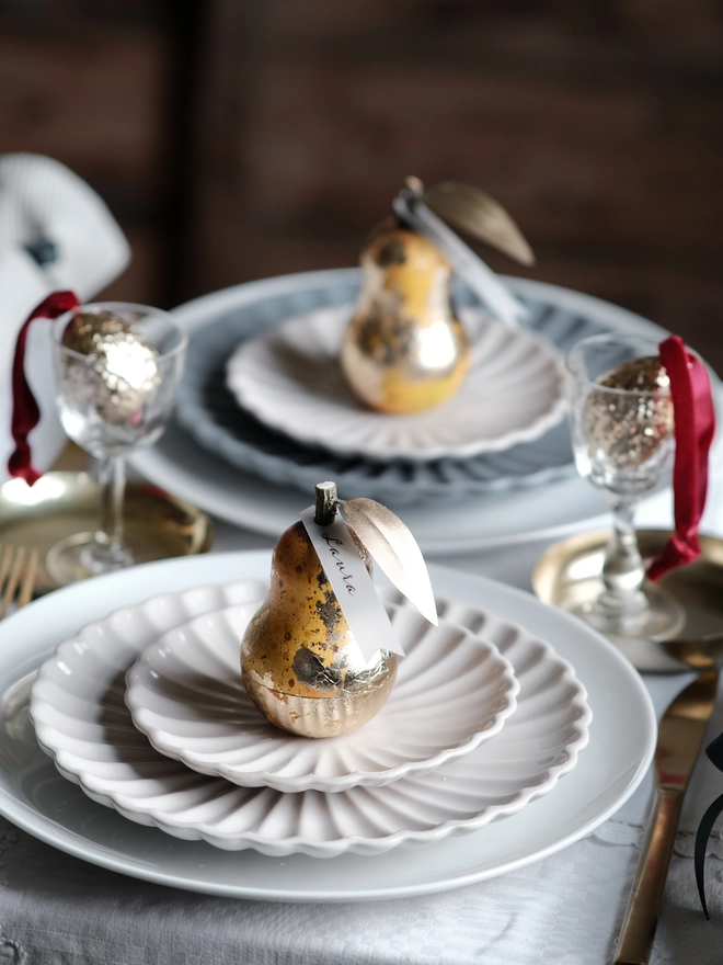 Gilded vintage-style Christmas pear ornament opened to reveal a small surprise inside, shown on a festive Christmas table setting with white plates and ribbon.