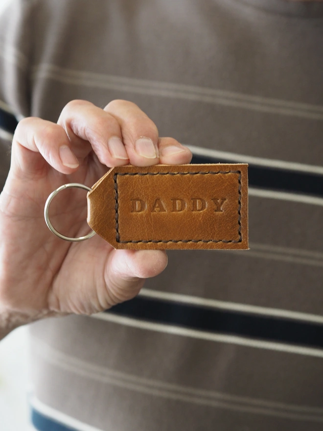 Personalised tan brown leather keyring. A classic tag shaped key ring held in a mans hand. The key ring has been stamped with the name DADDY.