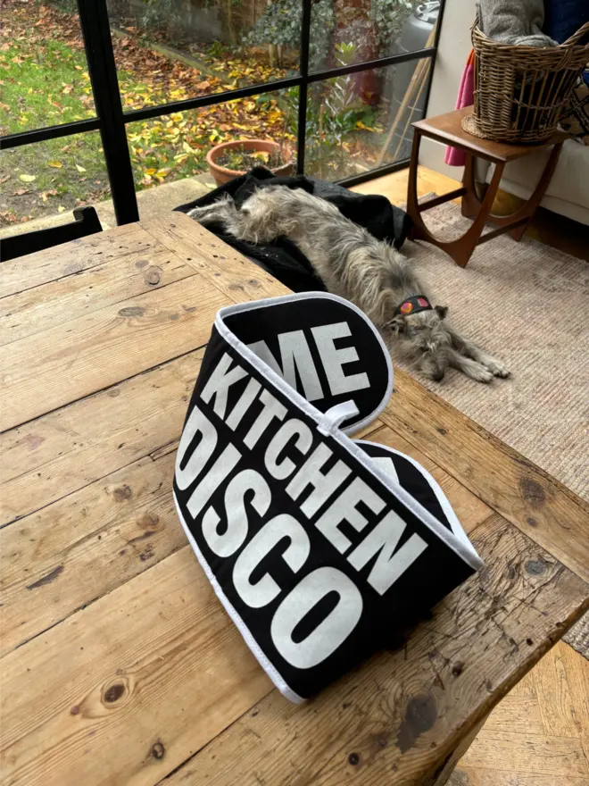 Black oven glove with white text that says KITCHEN DISCO on one side and DANCE WITH ME on the other side sat on a wooden table infront of a large window with a dig asleep on the floor in background