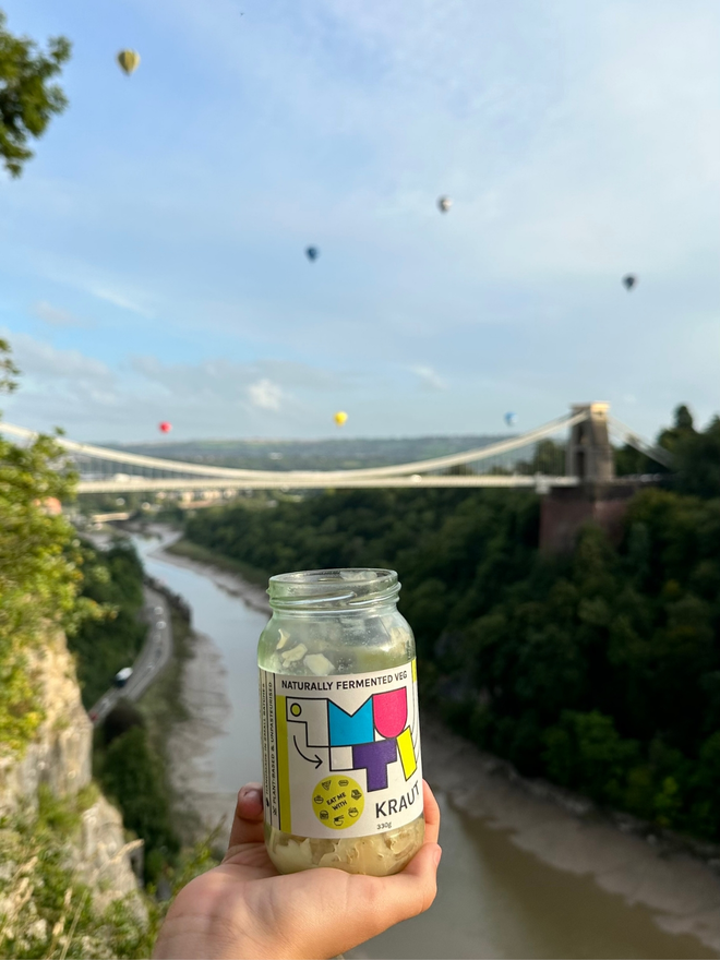 A Jar Of Muti Kraut Being Held Up To The Sky With Hot Air Balloons And The Clifton Suspension Bridge In The Background.