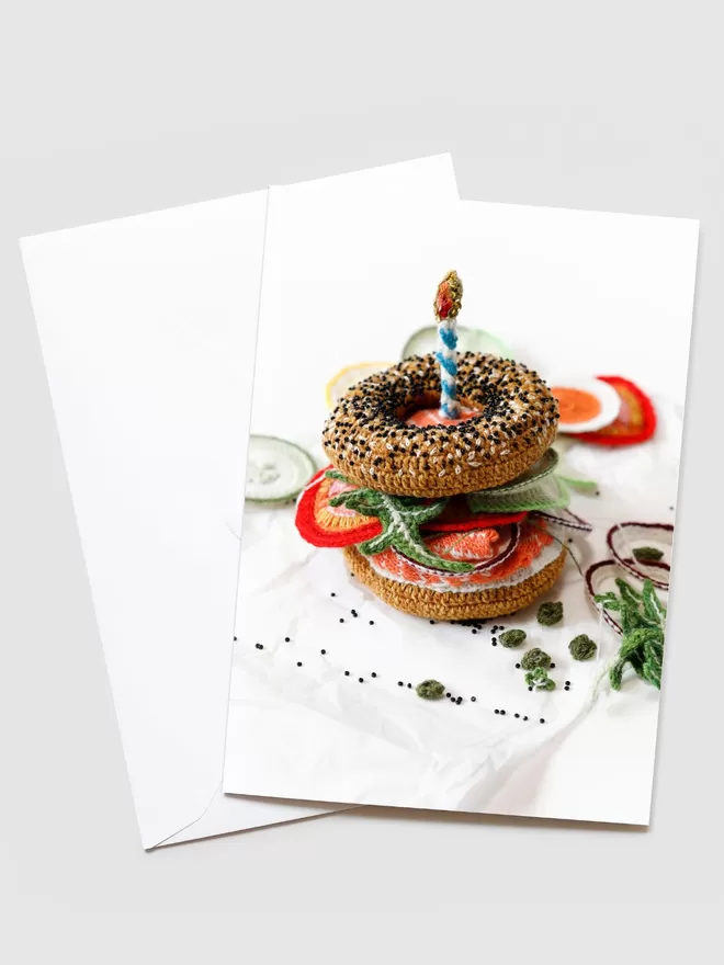 Kate Jenkins crotched Birthday Bagel seen on the front of a card with a white envelope behind. The bagel has a crotched candle on top with tomatoes and rocket peering out of the bagel.
