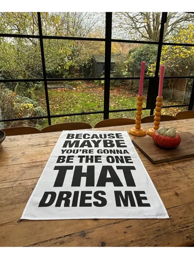 White tea towel with black text that says BECAUSE MAYBE YOU'RE GONNA BE THE ONE THAT DRIES ME laying on wooden table with chopping board, a pumpkin and yellow candlesticks with pink candles on table in front of large window