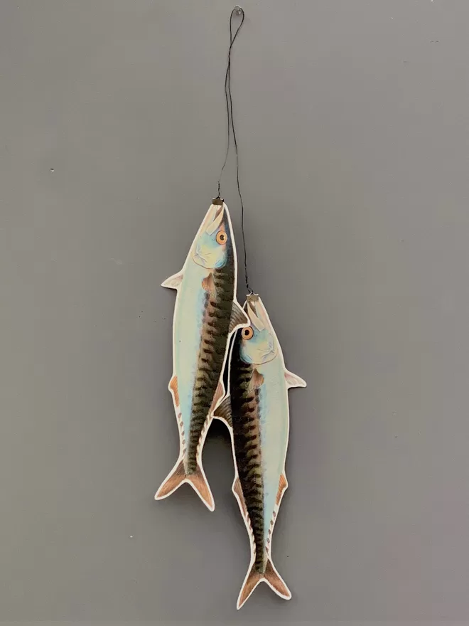 A sculptural wall hanging of two paper cut mackerel fish against a grey painted wall