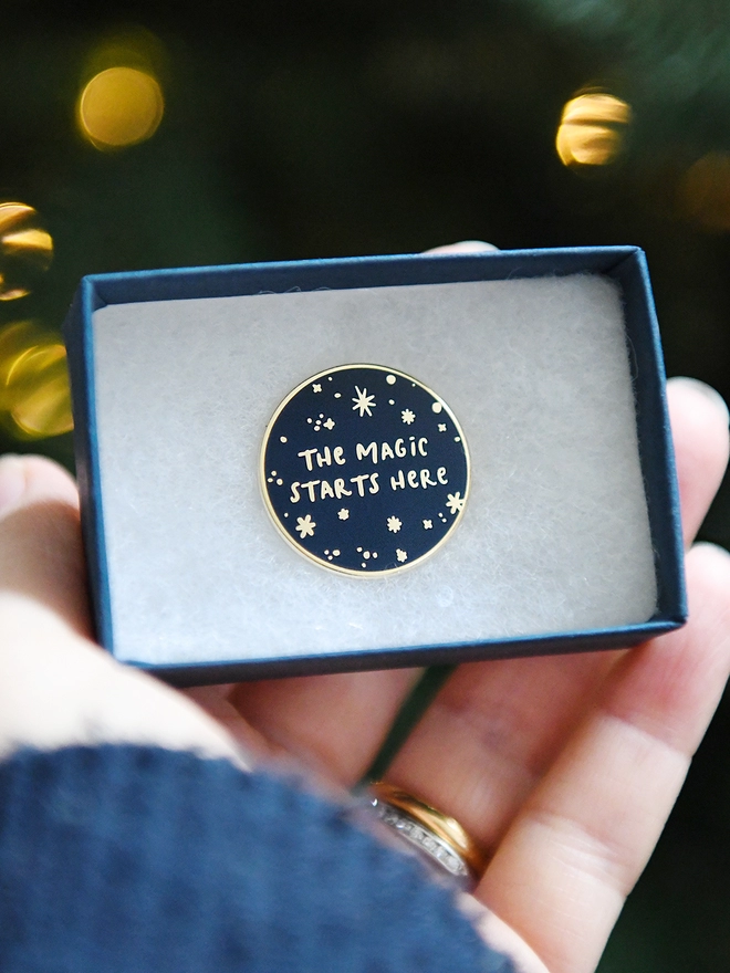 A navy blue and gold enamel pin badges with a starry design and the words "The magic starts here" is in an open gift box.