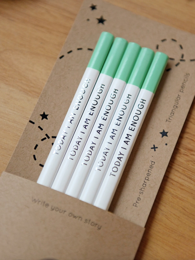 Five white pencils with green tips and the words Today I Am Enough written along the side, sit within cardboard packaging on a wooden desk with stationery items.