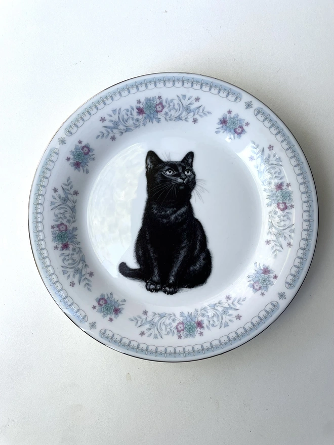a vintage plate with an ornate border, in the centre of the plate is a vintage style black and white illustration of a black witch's cat