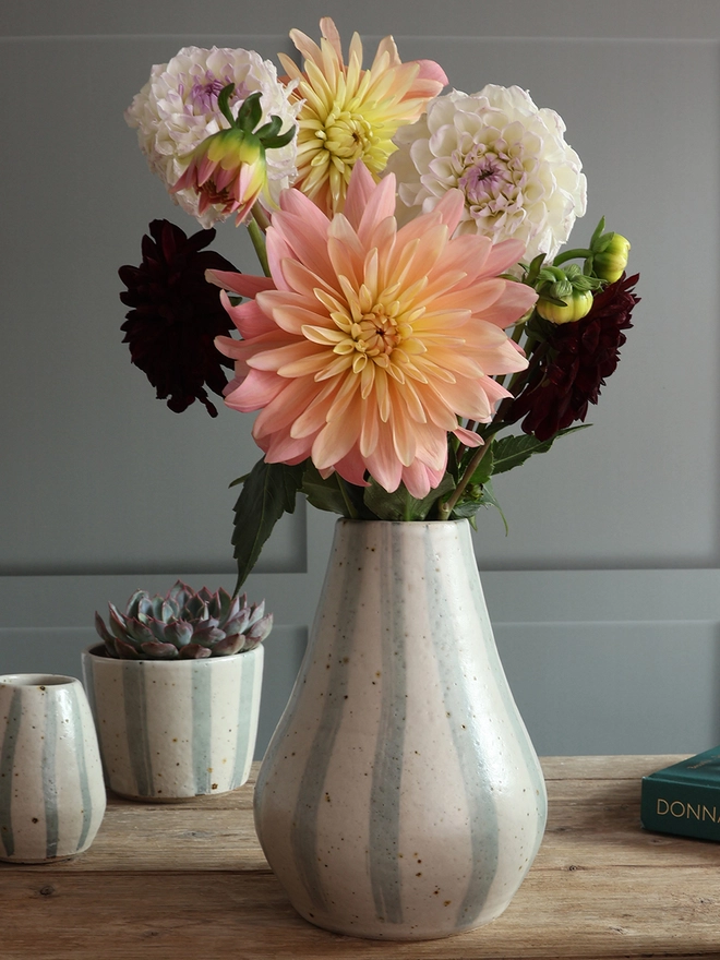 Handmade Ceramic Stoneware Rounded Vase in Blue and White Stripe filled with a bouquet of pink, yellow, white and red flowers. A small vase and plant pot holding a succulent plant sit behind the vase. 