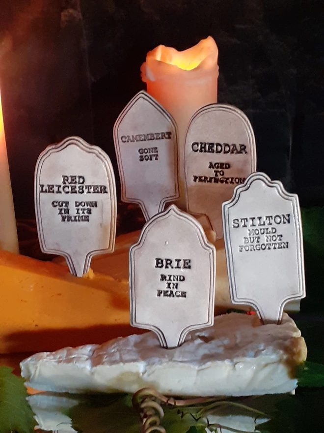 Five pale ceramic gravestone cheese markers arranged in Cheddar, Brie, Camembert, Red Leicester and Stilton, displayed among cheeses with glowing candles behind them.