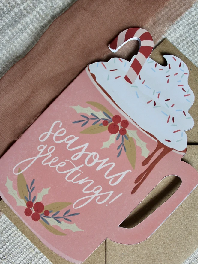 season's greetings christmas hot chocolate card. christmas card shaped like a pink mug with whipped cream on top.