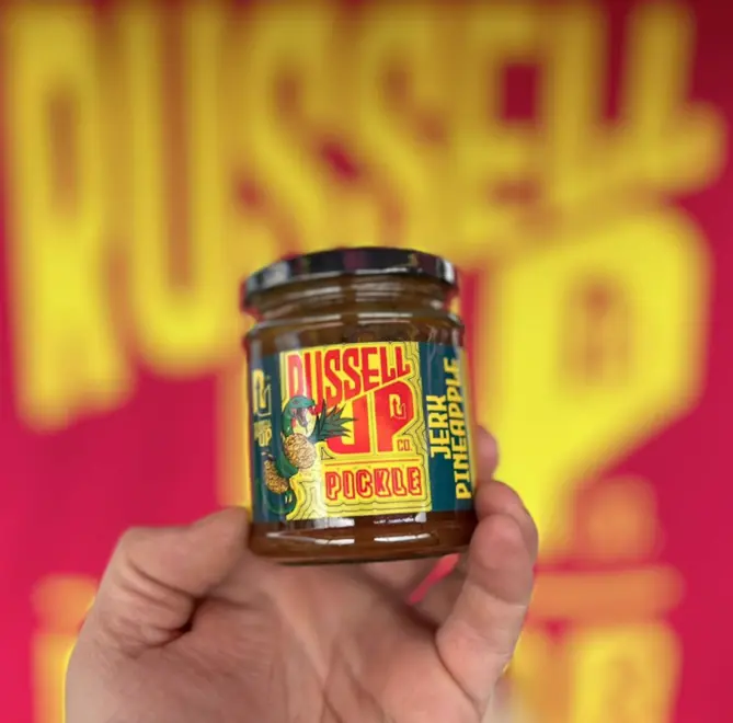 hand holding a jar of jerk pineapple jam in front of a blurred pink and yellow background reading "Russell Up"