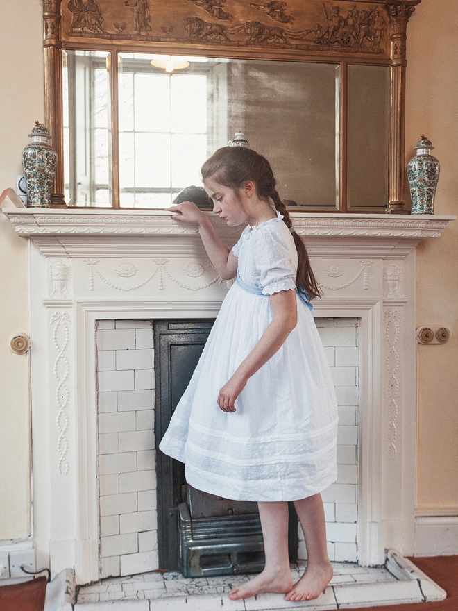 A girl in a white dress with a blue sash stands next to a fireplace