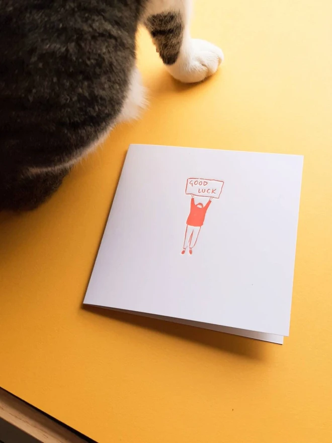 'Good Luck' Letterpress Handprinted Greetings Card, a white good luck card with an orange design against a yellow background. It is accompanied by a cat. 