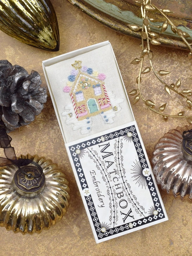 A delicious embroidered gingerbread house covered in sweets, inside a Matchbox displayed on a gold background with glass baubles.