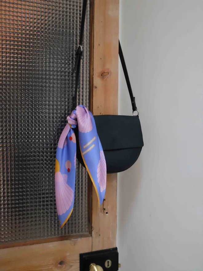 'Sea Shells - Blue and Pink' Silk Scarf tied around a leather handbag hanging on the back of a wooden doorframe