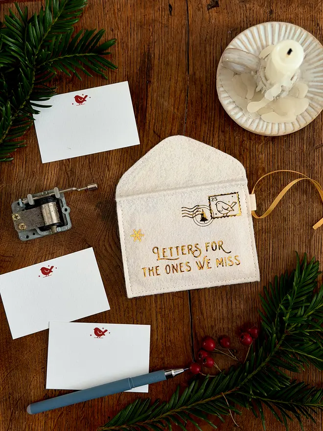 A handmade ivory felt envelope decoration with the words 'Letters for the ones we miss' in gold lays on a wooden table. Several robin notecards lay beside it.