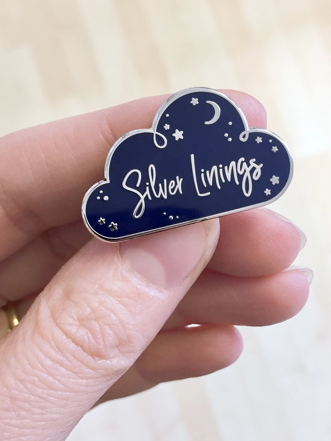 A navy blue and silver enamel pin badge in the shape of a cloud with a starry design and the words "Silver Linings" is being held in a hand.