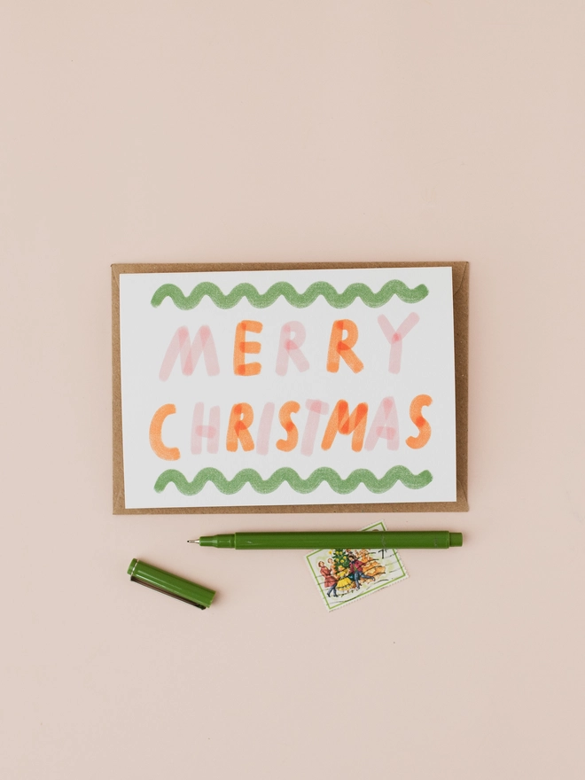 Christmas card featuring the words Merry Christmas in pink and red lettering and a green squiggly border