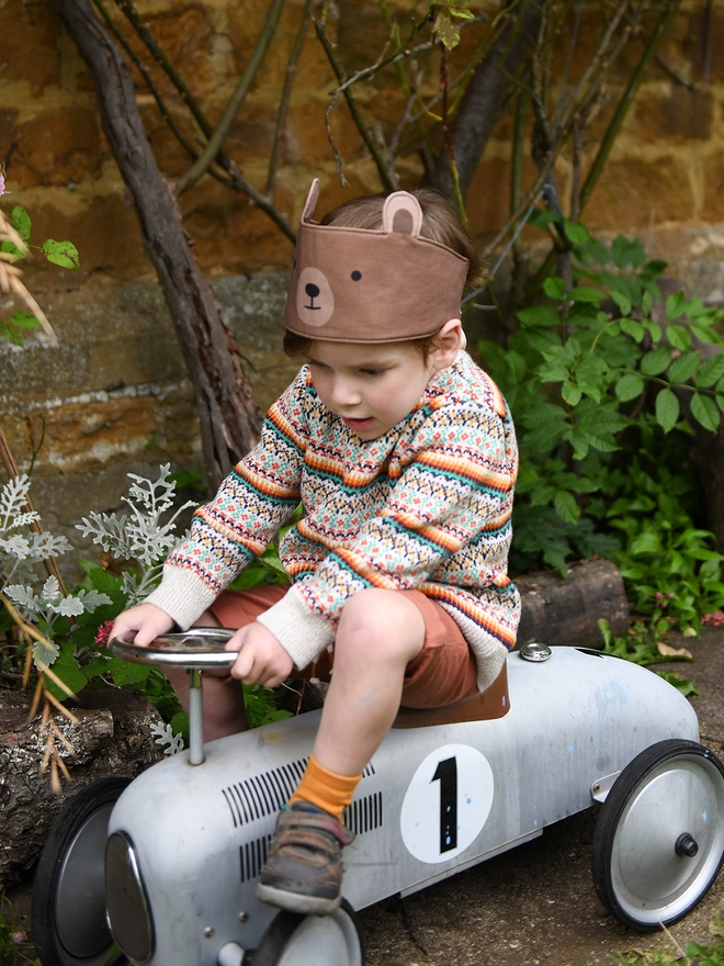 A young child wearing a knitted jumper and a bear dress up fabric crown sits on a ride on vintage car in the garden.