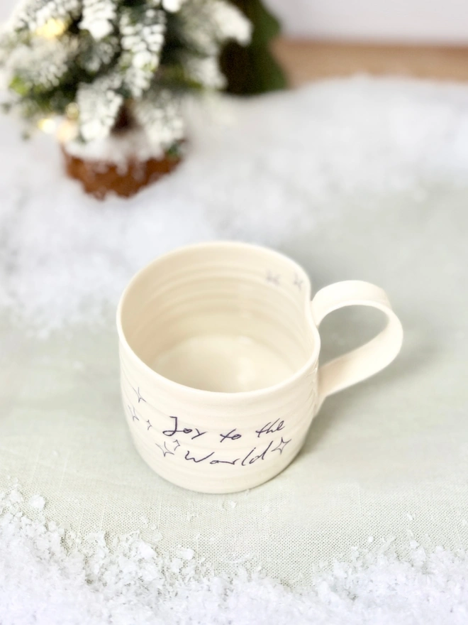 cream hand thrown porcelain mug with the words 'Joy to the World' printed onto the side with pretty line drawn stars. Sitting on a tabletop with snow, a mini lit Christmas tree 
