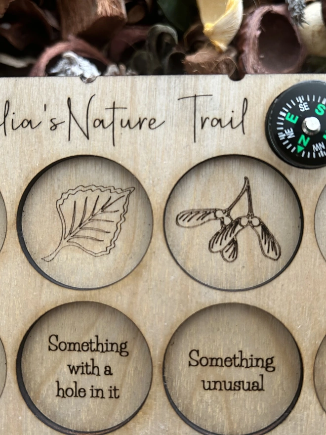 Close up of A personalised wooden nature scavenger hunt board displayed on an outdoor surface. The board is rectangular with rounded corners and engraved with 15 small collection slots held by clear bands. A custom name is engraved at the top, and a button compass with a wrist lanyard is attached for carrying. The wood has a smooth, light finish, and no items are placed in the slots; only the board is shown.