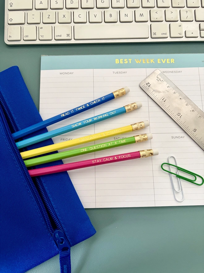 Exam essentials pencil set - Five colourful motivational exam pencils with gold foil lettering are styled on a pastel desk setup with stationery and a weekly planner.