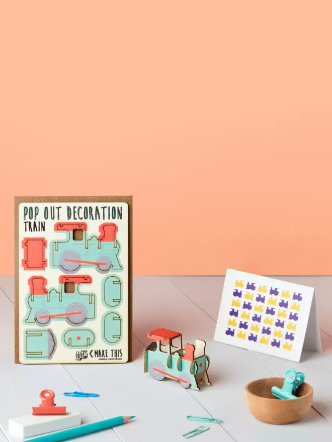 Pop out train decoration and train pattern greeting card and brown kraft envelope on top of a wooden desk in front of a peach-coloured background