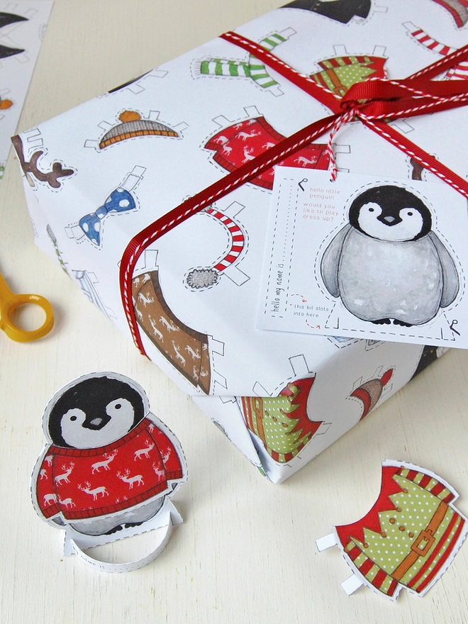 A gift wrapped in 'dress up a penguin' paper doll Christmas wrapping paper is on a white wooden table.