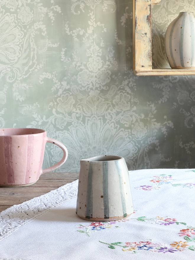 Handmade Ceramic Stoneware Small Jug in Blue and White Stripe sat on a wooden surface covered in a vintage floral tablecloth. A handmade ceramic mug with a pink stripe design sits behind the jug. 